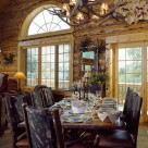 Custom wood dining table with bark on edges and bark on log dining chairs with antler chandelier above in handcrafted log home with french doors and sunrise grid arched window.