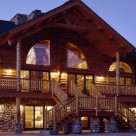 Twilight photo of amazing log duplex set on massive river rock faced lower level with grand entry framed by log pillars and log truss. Radius top windows backlit showing interior of homes.