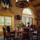 Large wood dining table with cozy dining chairs in Log home with french doors, wagon wheel chandelier and sunrise grid half circle window above door.