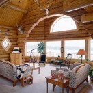 Interior log home living room with massive log arch framing bay windows with arch window above. Upholstered sofa and loveseat with end table sit on white carpet.
