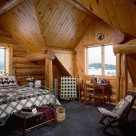 Loft bedroom in handcrafted log home with dormer valleys accenting large window with small desk and quilt on rack in dormer.