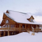 Exterior of custom log home covered in snow with bay windowed front, gable dormer and covered porch with log rails.