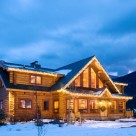 Twilight photo of custom log home with steep center gable filled with glass, shed dormers on either side and bright sunroom in front.