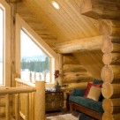 Cozy nook in loft of handcrafted log home with trapezoid window showing view of forest beyond.