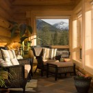 Sunroom in handcrafted log home with cozy chairs and ottoman with views of mountains and forest through lorge windows trimmed in pine.