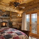 Master bedroom in luxury log home with stine fireplace in corner, exposed log beams in ceiling and views of Colorado mountains through french doors.