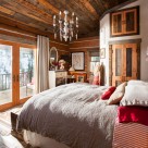 Bedroom of chink style log cabin with cozy bed, reclaimed wood ceiling, crystal chandelier and view to snowy forest through french doors.