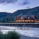 Twilight photo of magnificent handcrafted log home with river in foreground and Colorado mountains beyond.