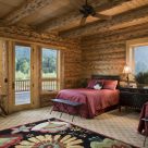Photo of handcrafted log home bedroom with two queen beds with red comforters and views through large widows and glass door to river and forest.
